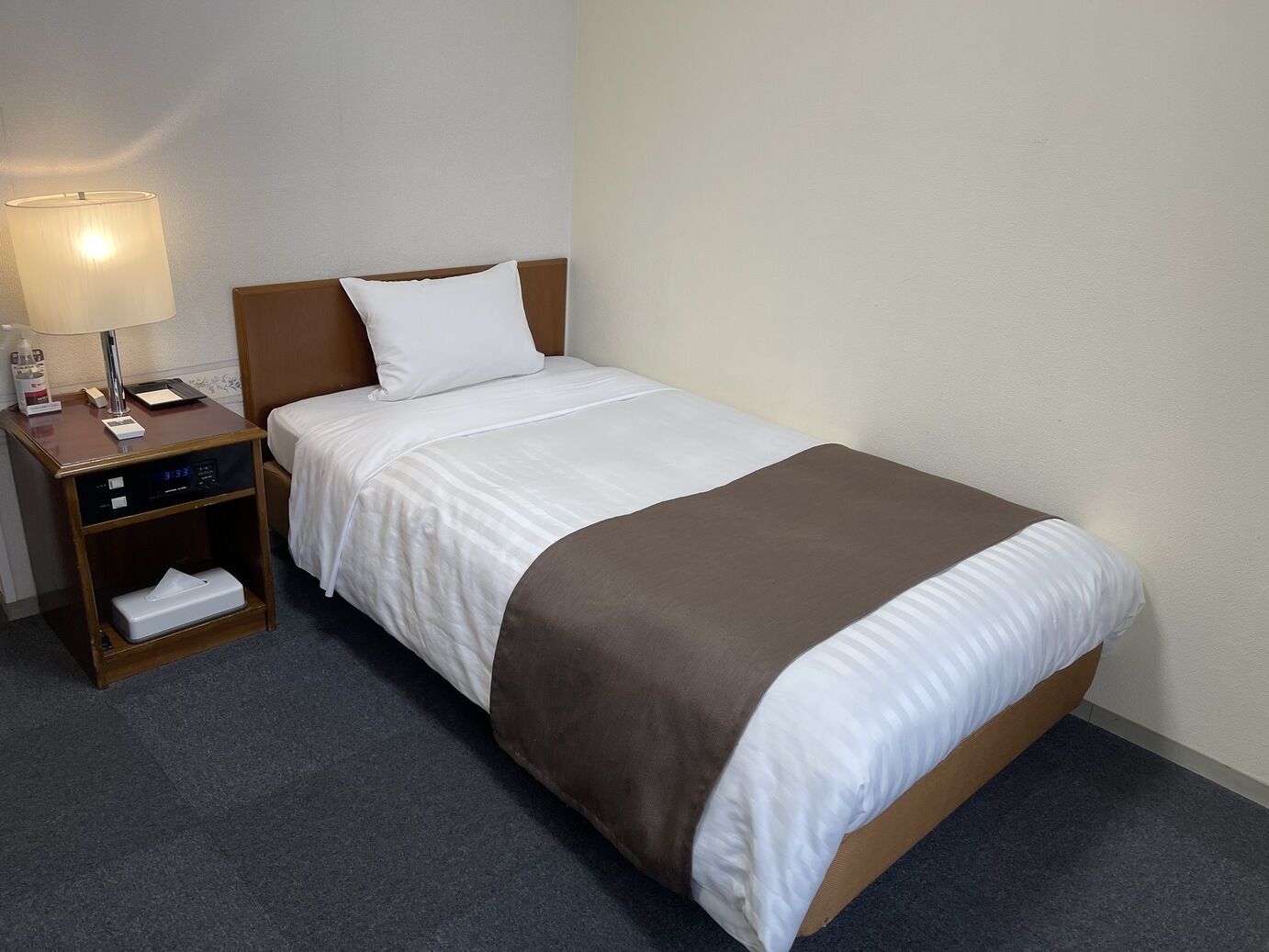 A hotel room with a bed, a nightstand with a lamp, a digital clock, and a tissue box.