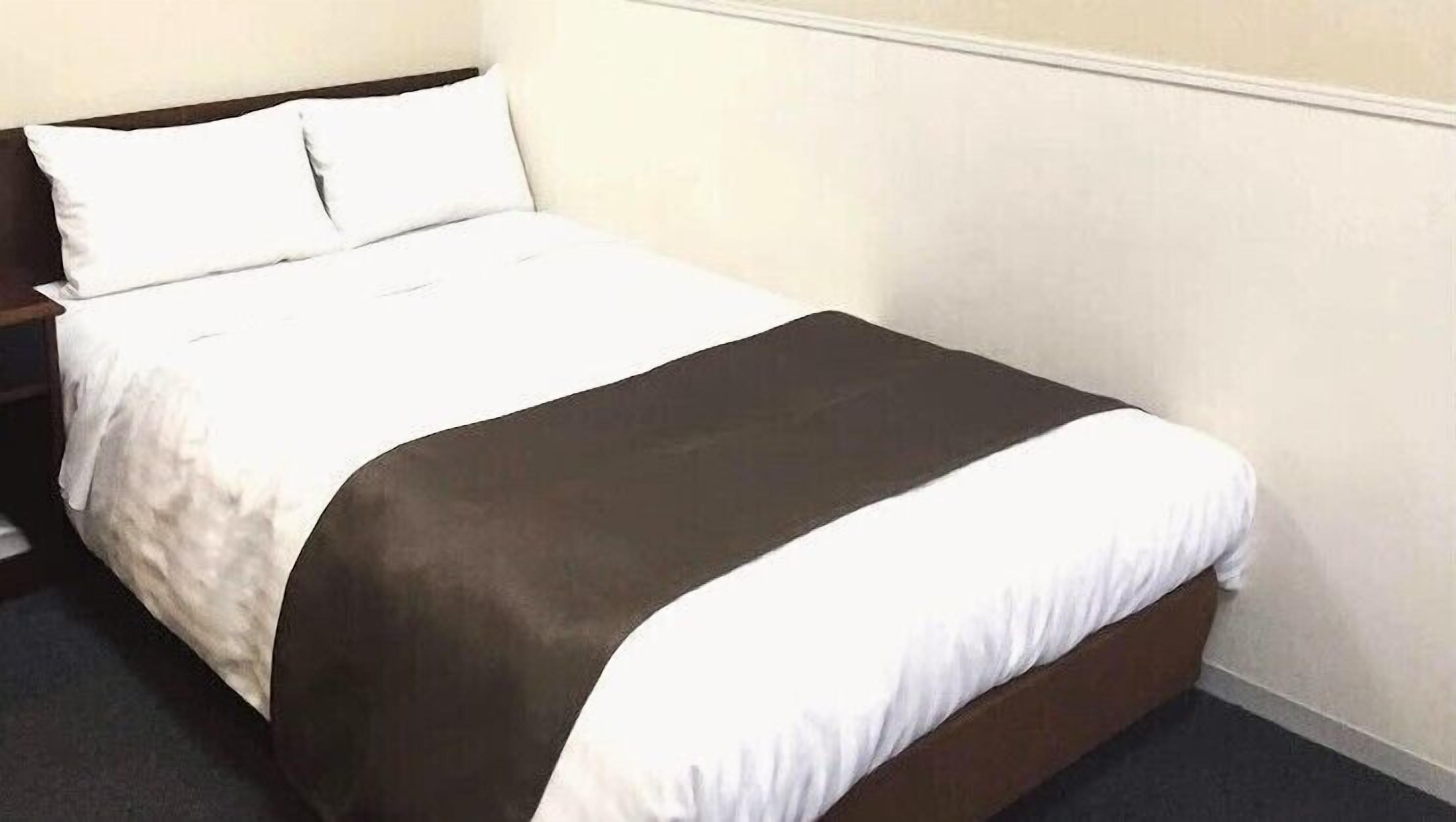 A neatly made bed with white and brown bedding, a wooden headboard, and a plain wall.