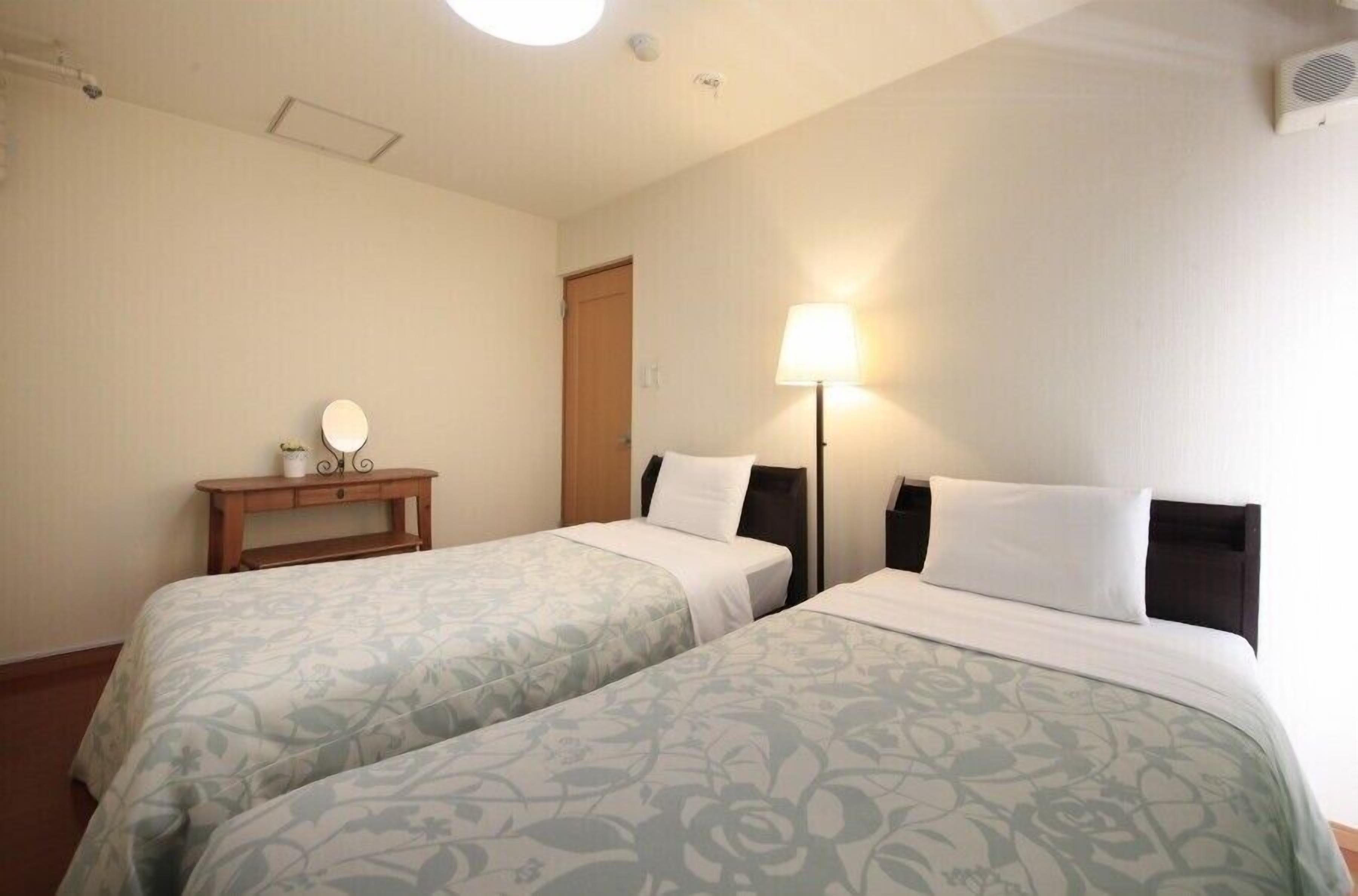 A hotel room with two beds, a bedside lamp, a small table with a mirror, and a door.