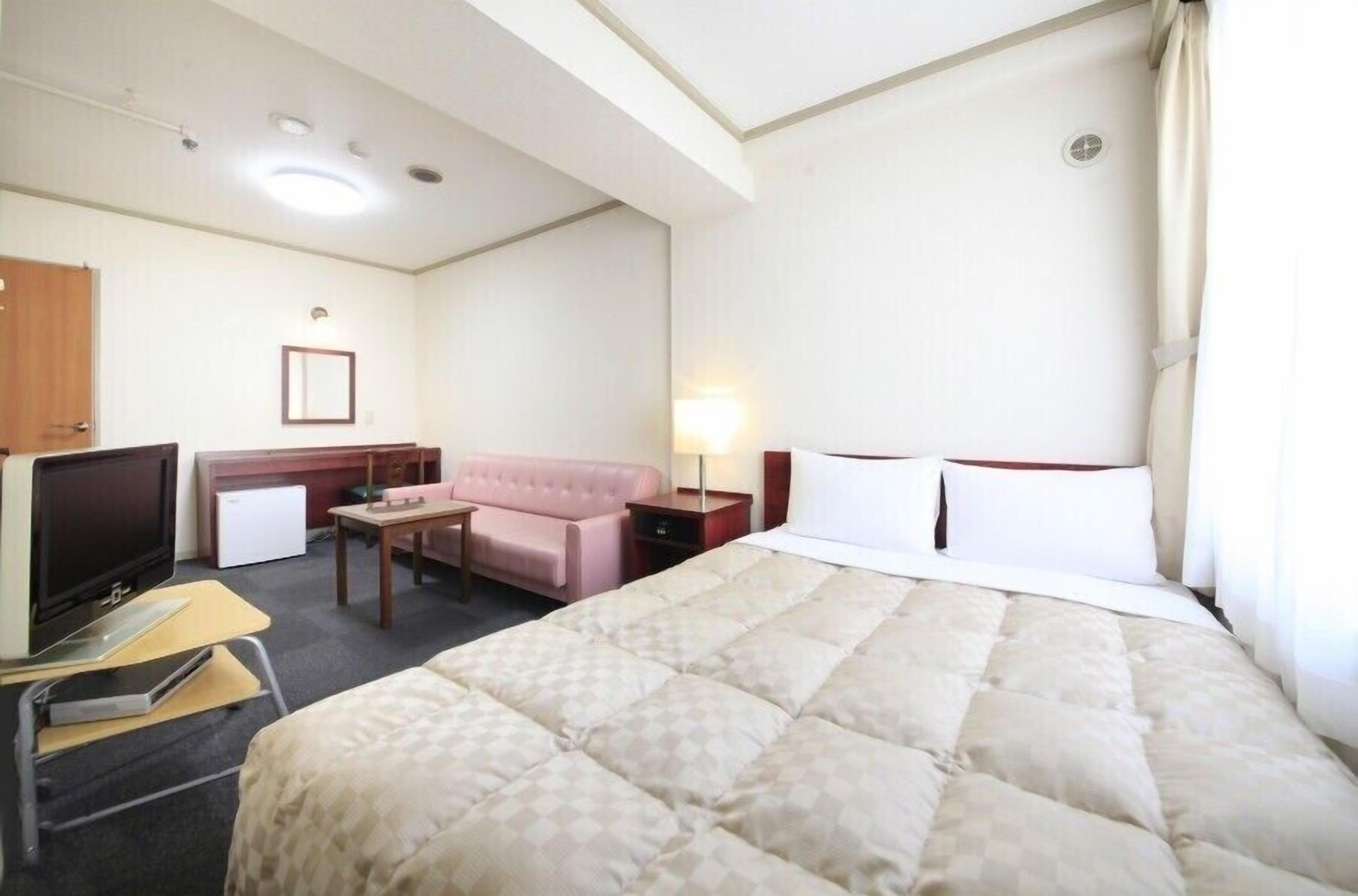 A hotel room with a bed, a desk with a computer, a TV, a sofa, and a lamp.