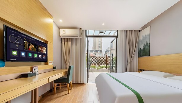 A hotel room with a bed, a desk with a computer, a TV, and a view of buildings through the window. - Greentree Inn Anqing Duxiu Avenue Greenland Metrop (Anqing)