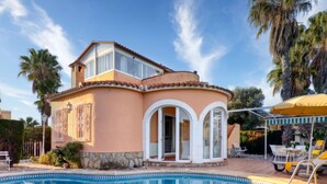 Ein zweigeschossiges Haus mit Swimmingpool, einer überdachten Terrasse mit Liegestühlen und Palmen.