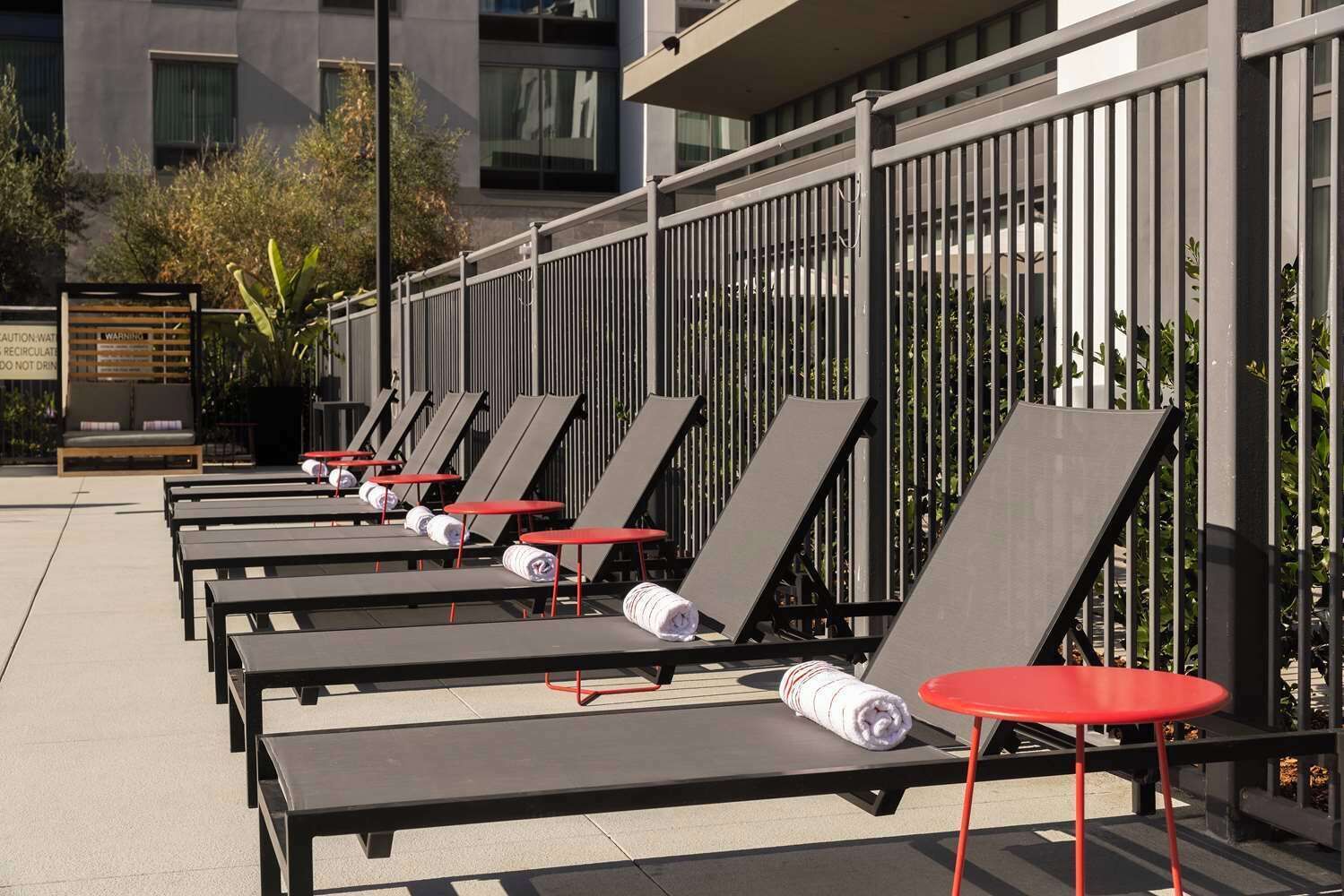 A row of sun loungers with red tables, a metal fence, and a building in the background.