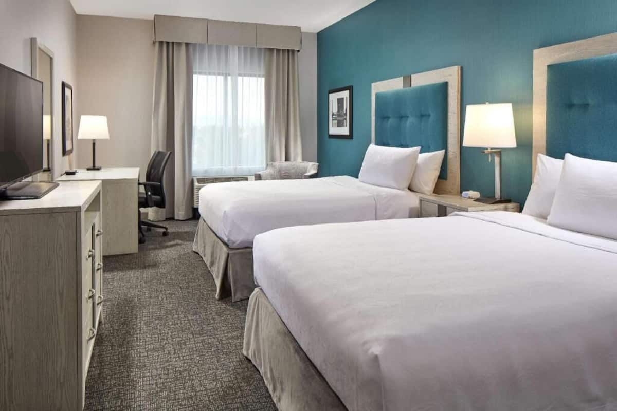 A hotel room with two beds, a desk, a chair, a TV, and a window with curtains.