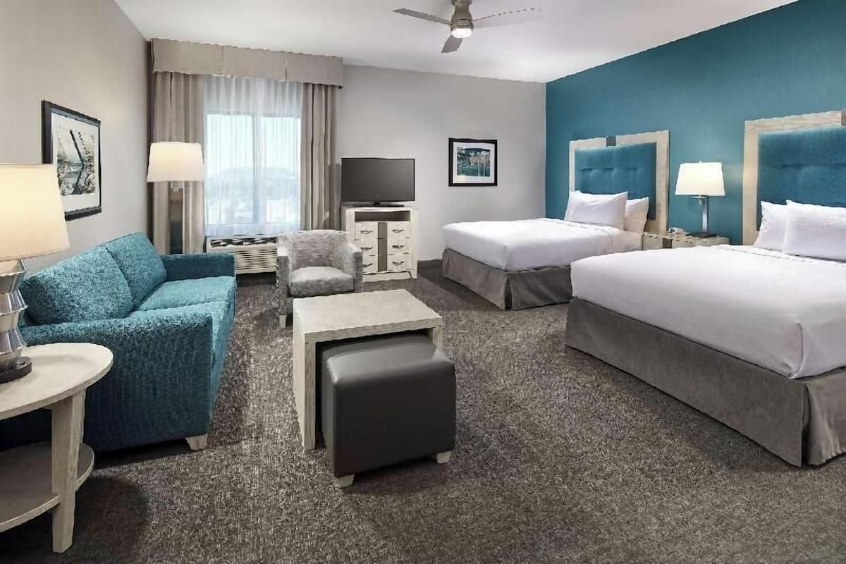 A hotel room with a sofa, two single beds, a TV, a desk, and a ceiling fan.
