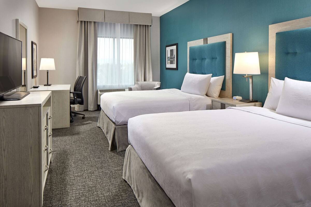 A hotel room with two beds, a desk, a chair, a television, and a window with curtains.