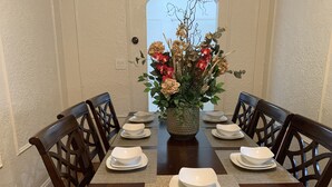 Ett matbord dukat med vita tallrikar och skålar, omgivet av mörka trästolar. En dekorativ vas med blommor står i mitten.