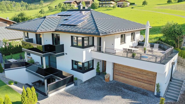 Una casa moderna de dos pisos con techo plano, ventanas amplias y un balcón con mobiliario exterior.
