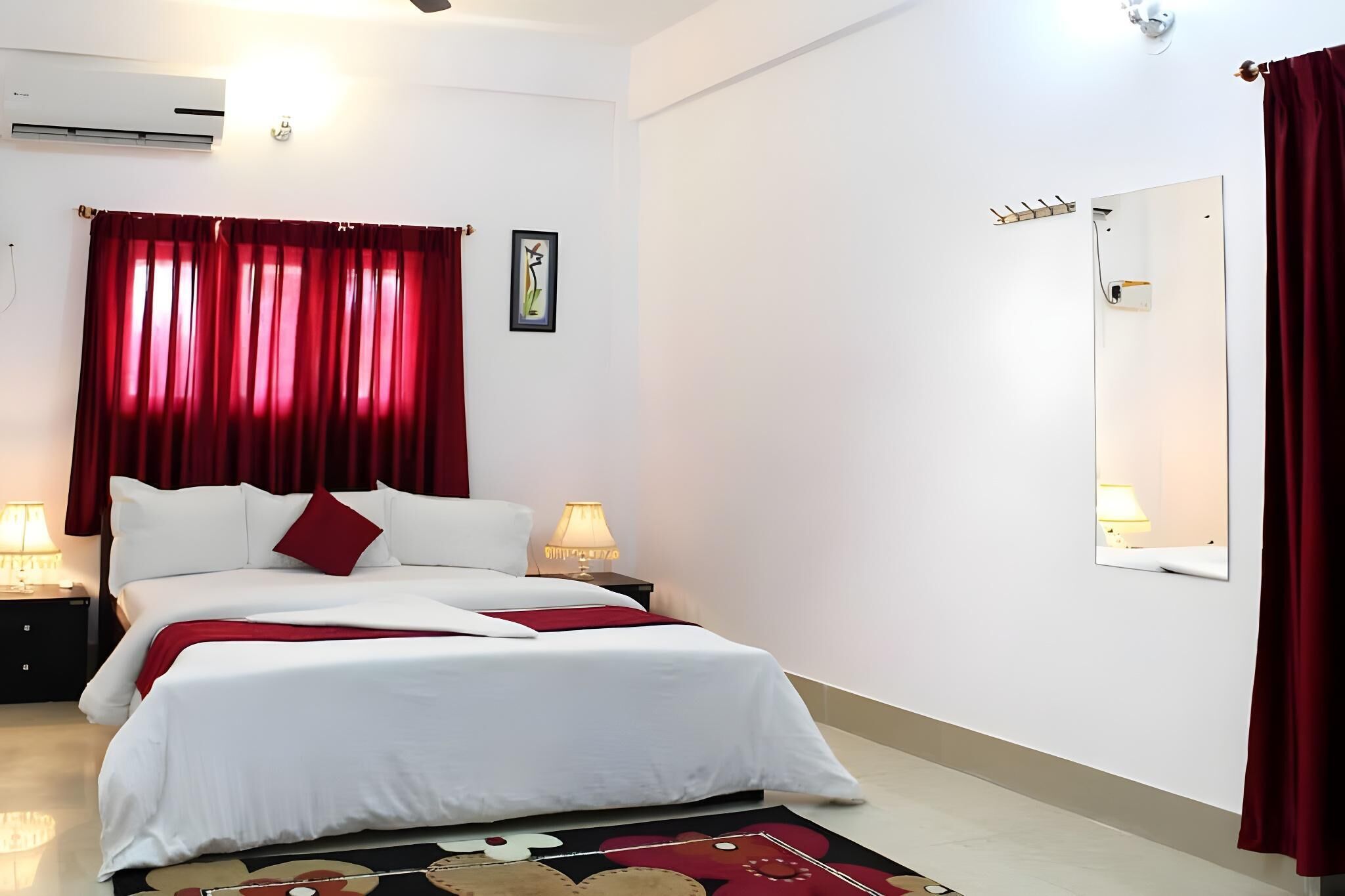 A hotel room with a bed, two bedside lamps, a red curtain, a wall-mounted mirror, and a small shelf.