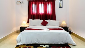 A hotel room with a bed, red curtains, two bedside lamps, an air conditioner, and a small picture on the wall. - The Shelter Hotel - Port Blair (Port Blair)