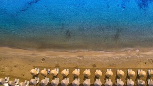 Vue aérienne d’une plage avec des rangées de transats et de parasols, un rivage sablonneux et une eau d’un bleu limpide.