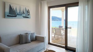 Una sala de estar con un sofá, un cuadro de veleros y un ventanal corredizo que da a una zona de estar exterior con vistas al mar.