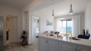 Una cocina moderna con una isla grande, armarios blancos e iluminación colgante. Se ve un comedor con vista al mar a través del ventanal.