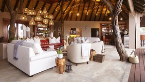 A spacious living area with a high ceiling and exposed wooden beams, furnished with white sofas and a red leather couch, surrounded by natural wood elements and hanging wicker lamps.