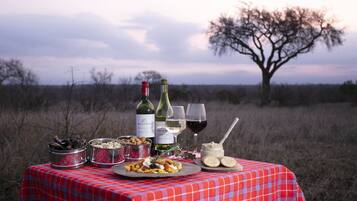 Un montaje para picnic con mantel a cuadros rojos, botellas de vino, vasos y recipientes de comida en un campo abierto con un árbol de fondo.