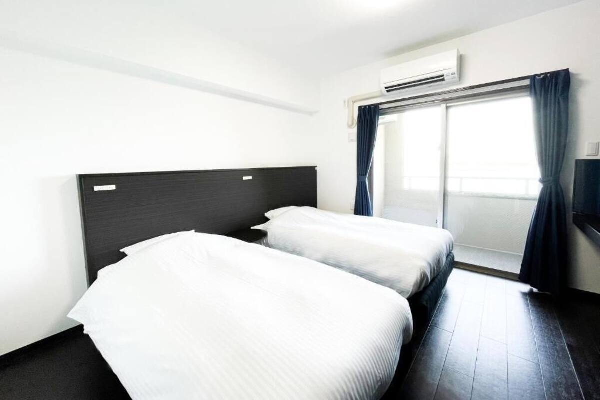 A hotel room with two single beds, a dark headboard, a large window with curtains, and an air conditioning unit.