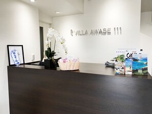 Ein Empfangsschalter mit dem Schild 'Villa AwaSe 111'. Es befindet sich ein gerahmtes Bild, eine Topforchidee und eine Korb mit Gegenständen auf dem Schalter.