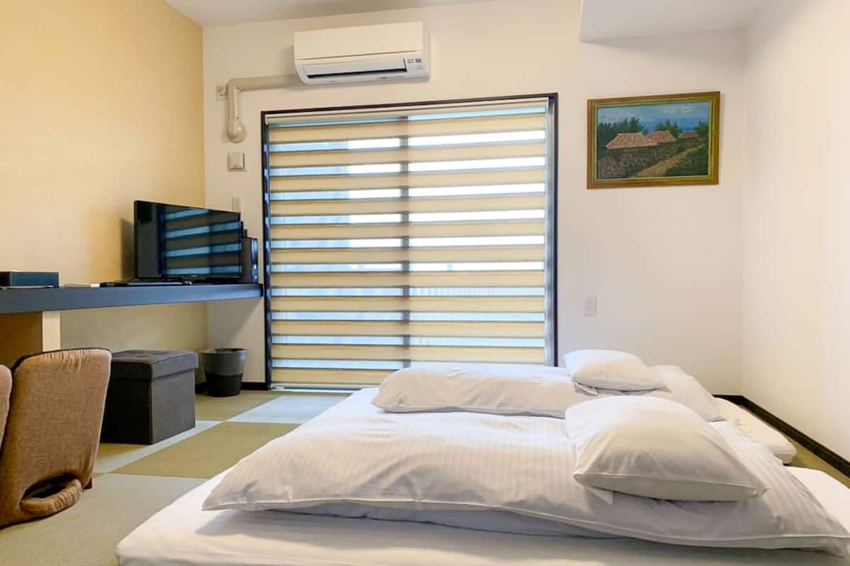 A hotel room with a bed, a desk, a chair, an air conditioner, and a window with blinds.