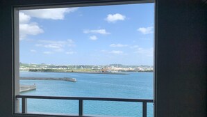 A view from a window overlooking a body of water with a distant cityscape, including buildings and a crane. - Villa Awase 111 (Okinawa)