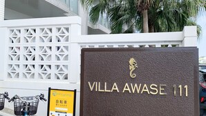 A sign for "Villa Awaase 111" with a modern building in the background, a palm tree, and a bicycle parked nearby. - Villa Awase 111 (Okinawa)