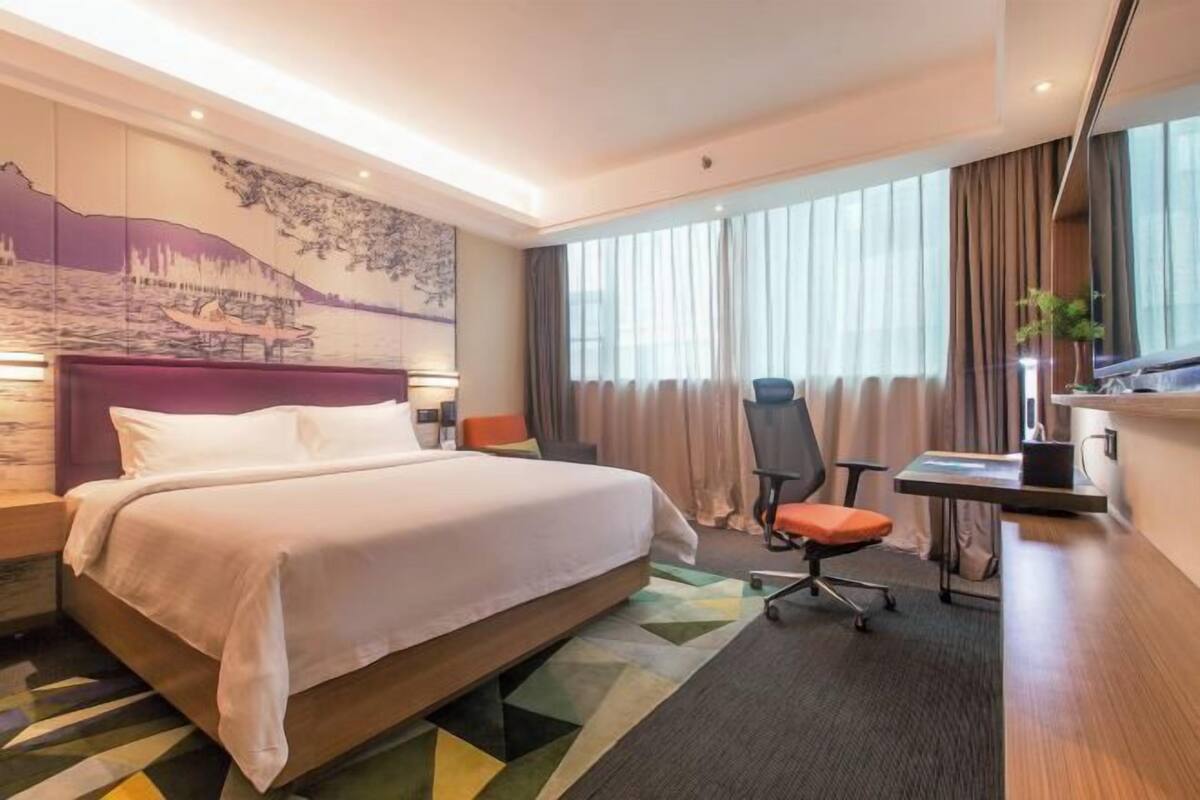 A hotel room with a large bed, a desk with a chair, a large window with curtains, and a wall mural.