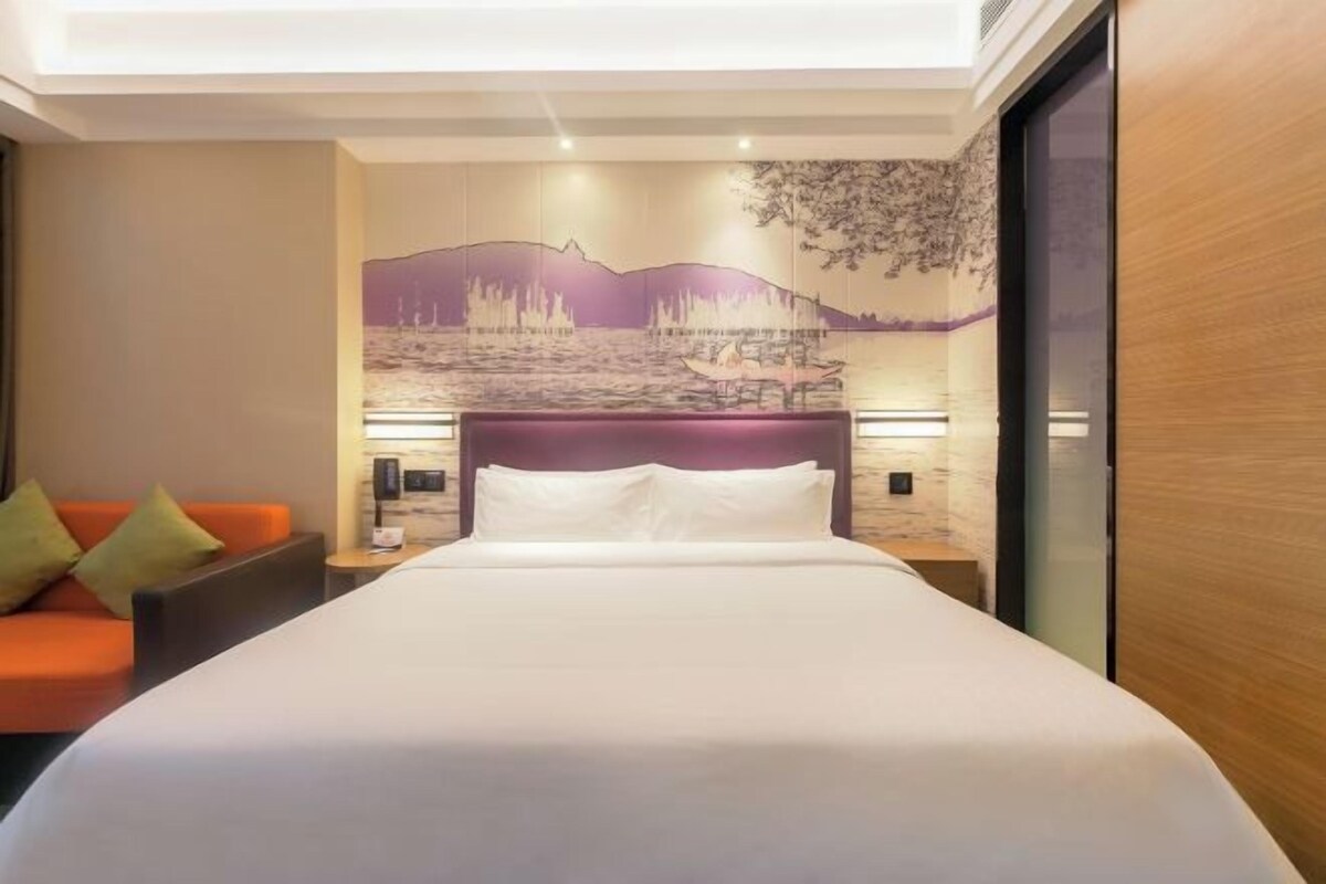 Una habitación de hotel con cama, mesita de noche, lámpara, un mural en la pared y un sofá naranja.