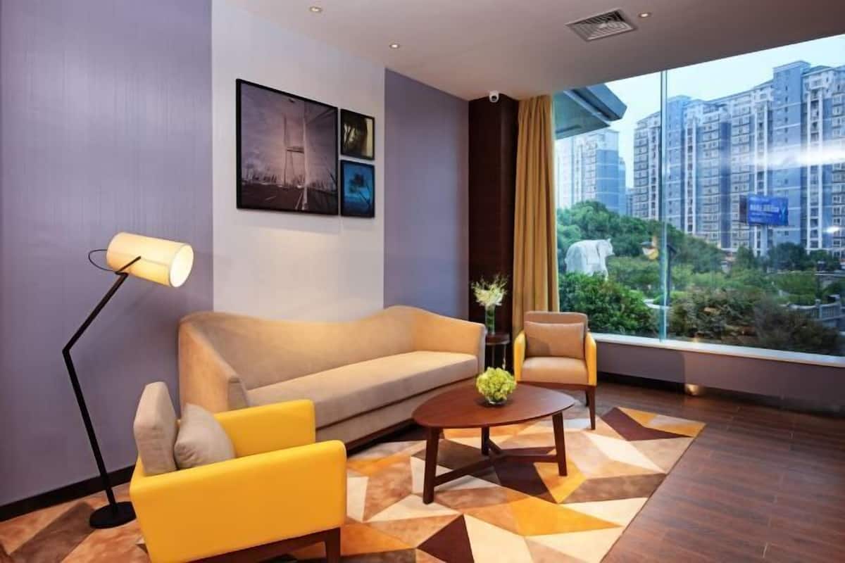 A modern hotel room with a sofa, armchair, and a coffee table. There are framed pictures on the wall and a window with a view of high-rise buildings.