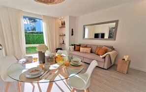 Ein modernes Wohnzimmer mit einem Glastisch, weißen Stühlen, einer Couch mit orangefarbenen Kissen und einem großen Spiegel an der Wand.