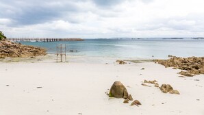 Uma praia com areia branca, pedras espalhadas e um píer que se estende em águas calmas.