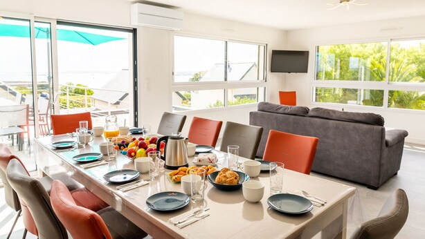 Uma área de jantar moderna com uma mesa comprida posta para o café da manhã, grandes janelas com vista para o exterior e um sofá cinza ao fundo.