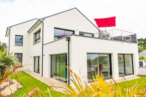 Une maison moderne à deux étages, dotée de grandes fenêtres en verre, d’un toit plat et d’un parasol rouge sur le niveau supérieur.