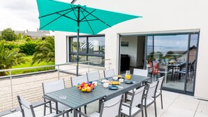 Uma área de refeições moderna ao ar livre, com mesa posta para quatro pessoas, um guarda-sol verde-água e vista para a vegetação e edifícios circundantes.