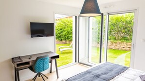 Quarto com cama, escrivaninha com cadeira, televisão na parede e porta de vidro deslizante para um jardim.