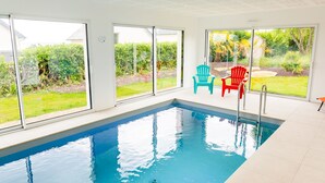 Piscina interna com paredes de vidro transparente, duas cadeiras e vista para um jardim.