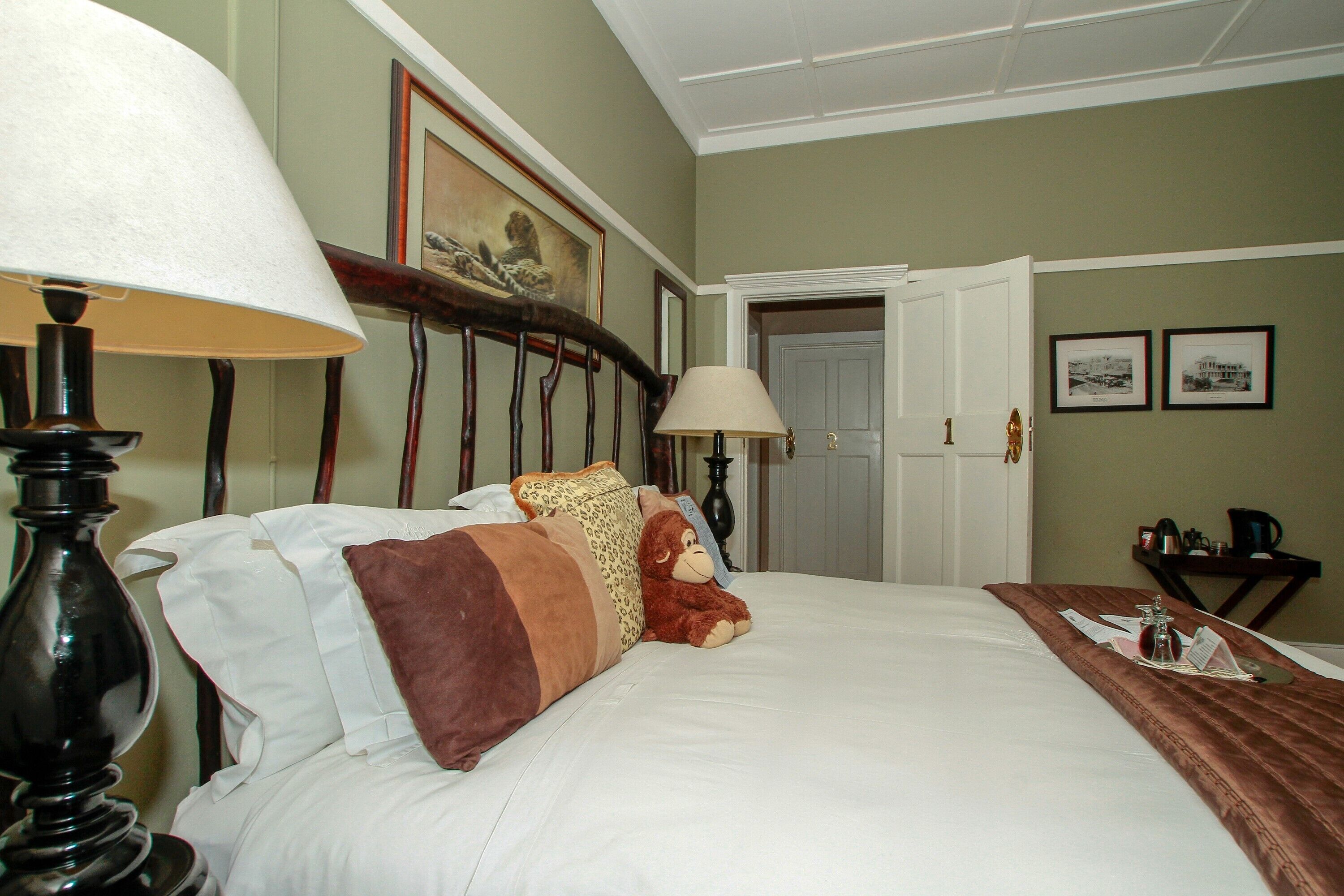 A hotel room with a bed, two lamps, a teddy bear, a brown throw blanket, and a small table with a tray.