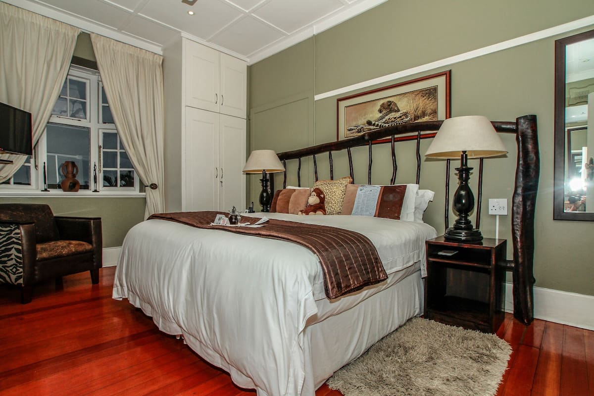 A bedroom with a large bed, two bedside tables with lamps, a chair, a window with curtains, and a painting on the wall.