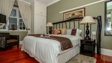A bedroom with a large bed, two bedside tables with lamps, a chair, a window with curtains, and a painting on the wall.