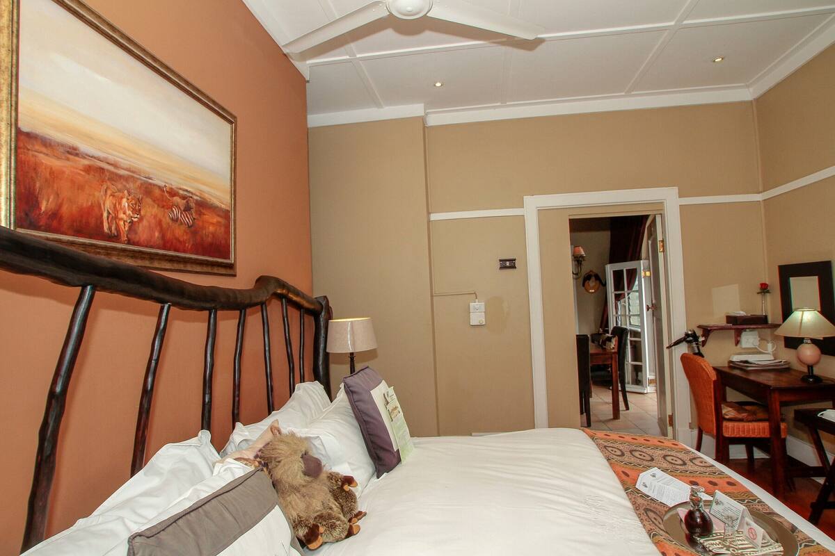 A hotel room with a bed, a desk, and a chair. There is a painting on the wall and a ceiling fan.