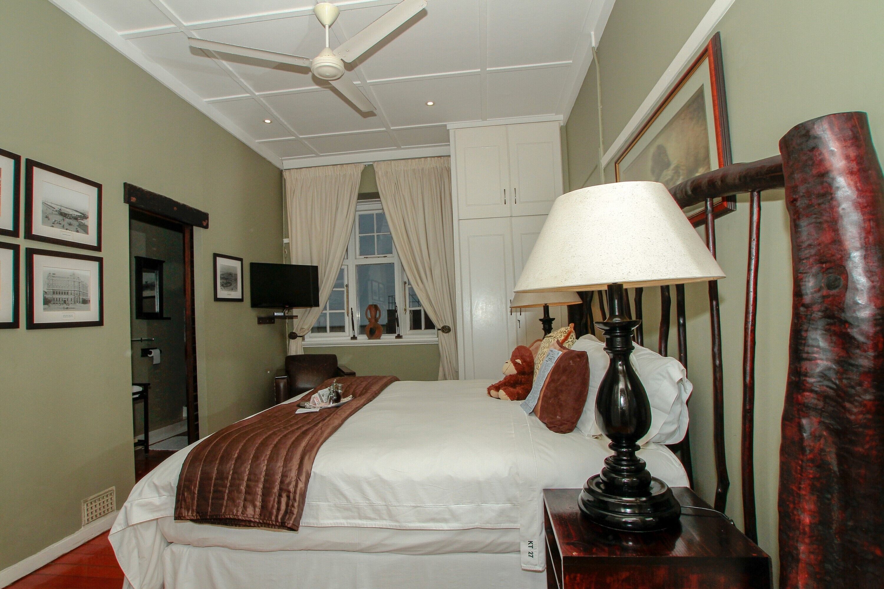 Une chambre à coucher bien rangée, avec un lit, une lampe de chevet, une commode, un miroir et une fenêtre avec des rideaux.