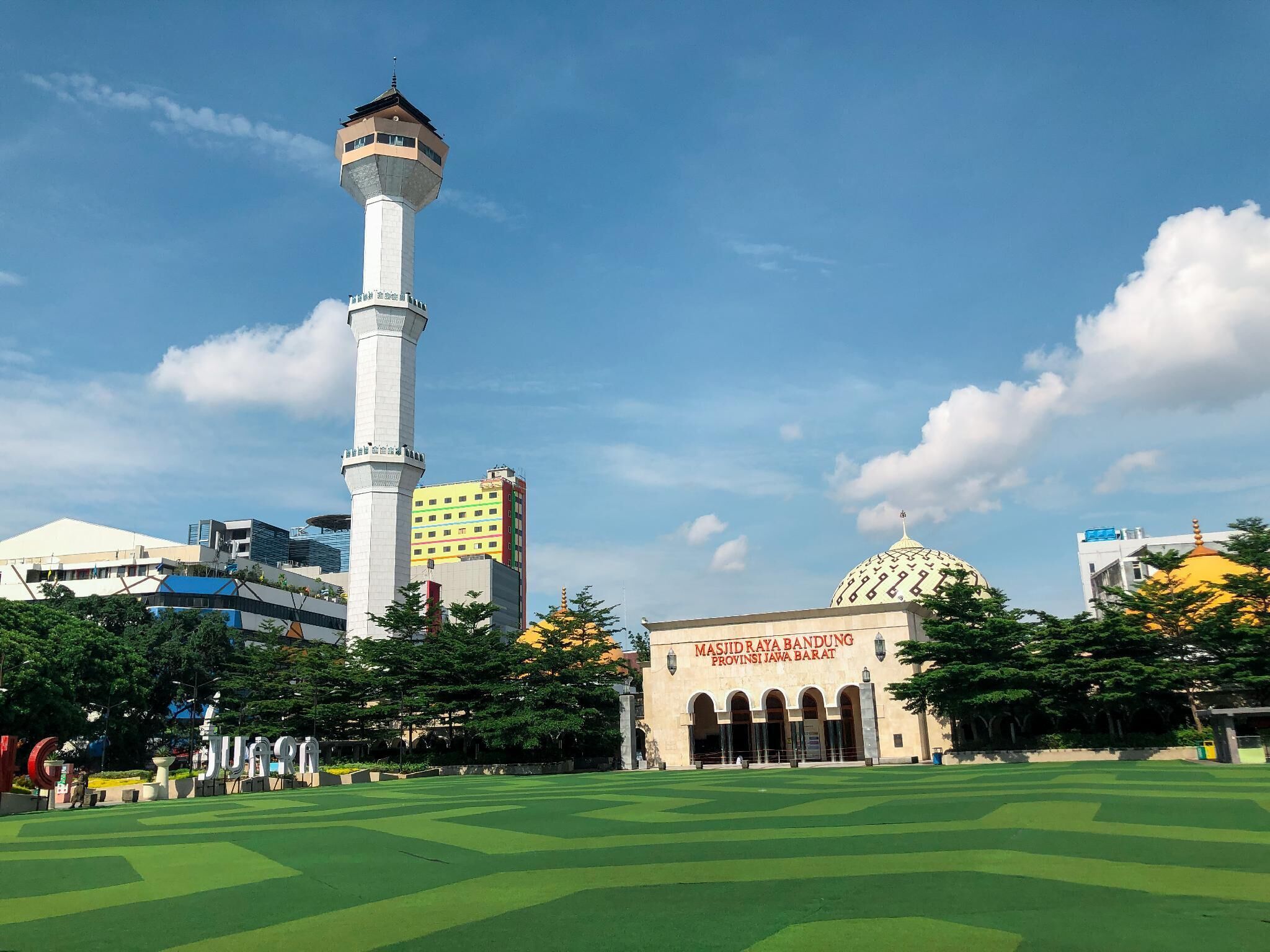A mosque with a tall minaret and a golden dome, surrounded by greenery and modern buildings.