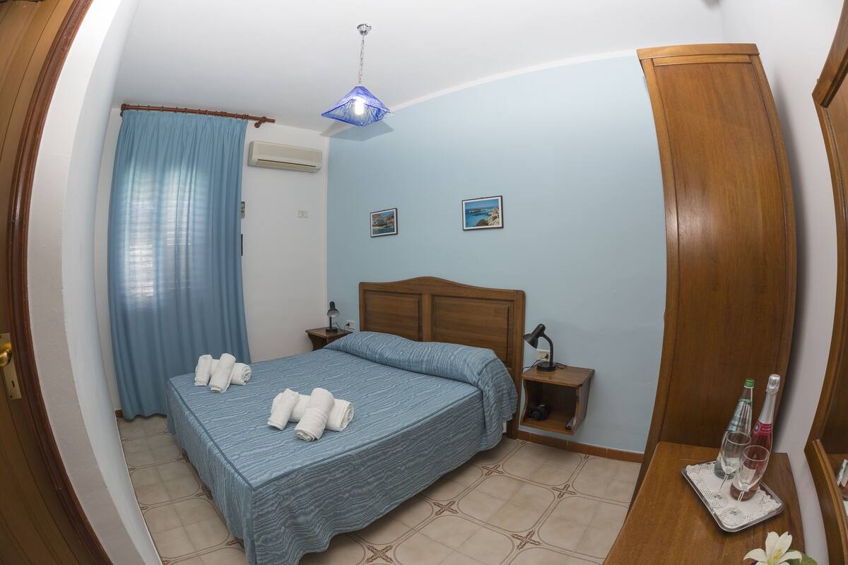 A bedroom with a bed, a nightstand, a bedside table, a lamp, a curtain, and a wall-mounted air conditioner.
