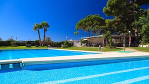 En klarblå pool med ett avskilt område för barn, omgiven av grönska och en byggnad med uteservering.