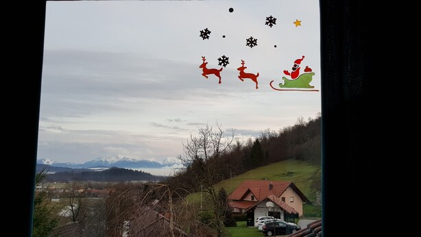 Landskab med et hus og parkerede biler, omgivet af bjerge og en skyet himmel.