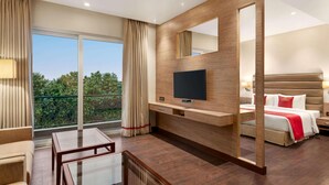 A hotel room with a large window, a flat-screen TV, a bed, a bedside table, and a seating area with a table and chairs. - Ramada By Wyndham Aligarh Gt Road (Aligarh)