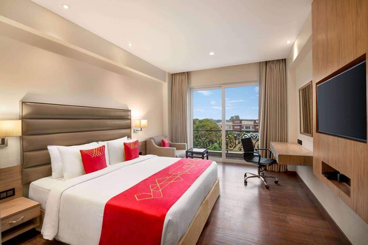 A hotel room with a large bed, a desk, a chair, a TV, and a balcony with a view.