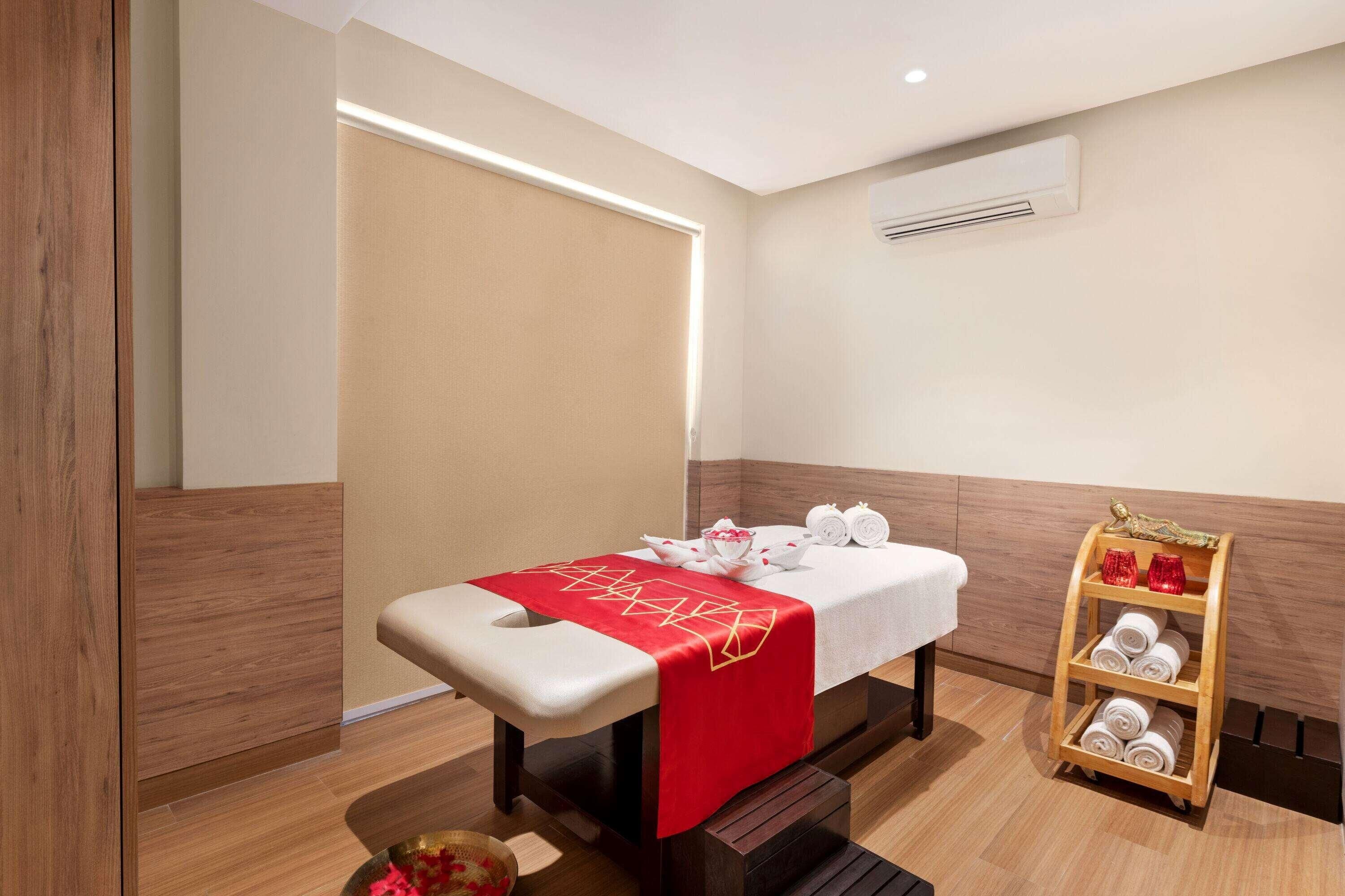 A massage room with a wooden bench, a massage table, a small shelf with towels, and a wall-mounted air conditioner.