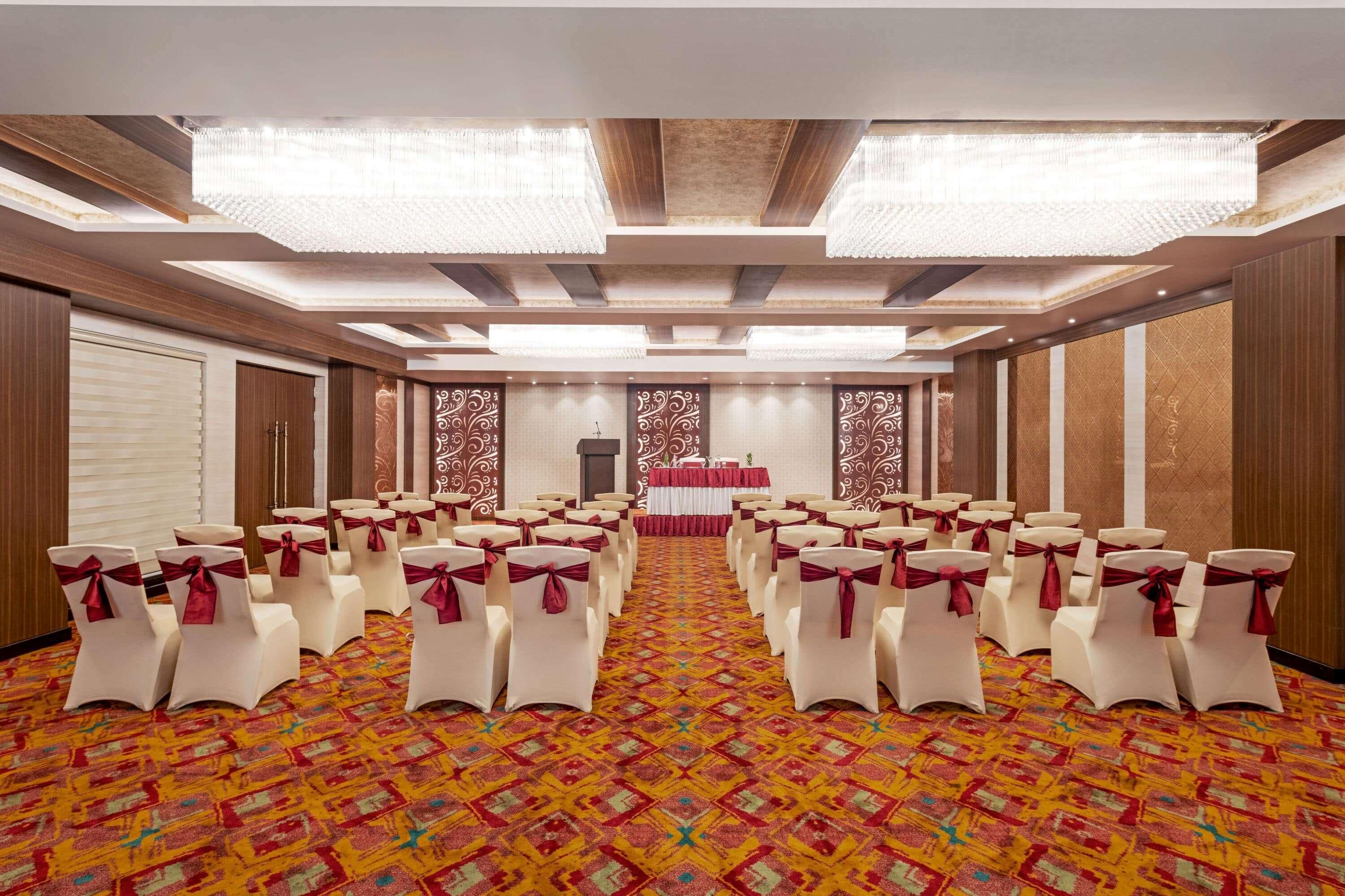 A conference room with rows of white chairs with red sashes, a patterned carpet, and a stage with a red backdrop.