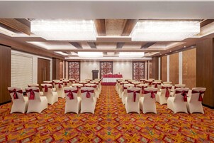 Un salón de conferencias con filas de sillas blancas con cintas rojas, alfombra estampada y un escenario con fondo rojo.