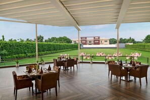An outdoor dining area with tables and chairs, a view of a golf course, and a building in the background. - Ramada By Wyndham Aligarh Gt Road (Aligarh)