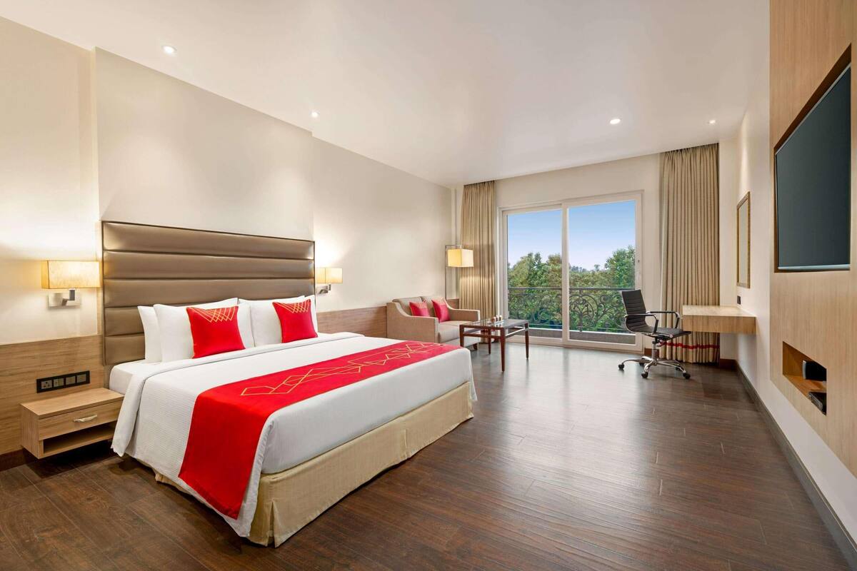 A hotel room with a large bed, a desk, a chair, a TV, and a balcony with a view.